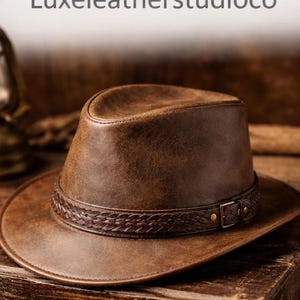 May include: A brown leather hat with a braided band and a buckle. The hat has a wide brim and a structured crown. The text "Luxeleatherstudioco" is at the top of the image. The hat is sitting on a wooden surface.