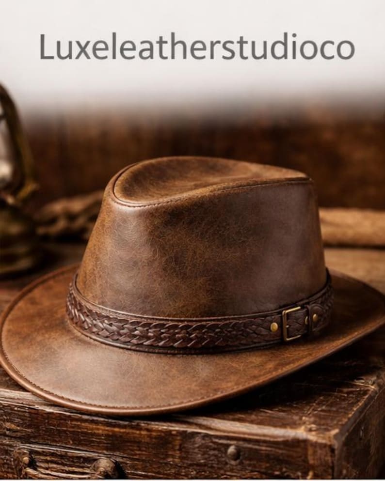 May include: A brown leather hat with a braided band and a buckle. The hat has a wide brim and a structured crown. The text "Luxeleatherstudioco" is at the top of the image. The hat is sitting on a wooden surface.