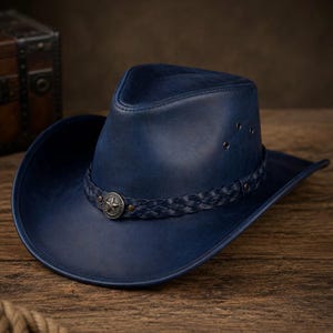 May include: A deep blue leather cowboy hat featuring a braided leather hatband and a circular silver-toned metal emblem with a star design at the front. The hat has a pinched crown, a curved brim, and small metal ventilation eyelets on the side.