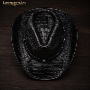 K&ouml;nnte beinhalten: Ein schwarzer Cowboyhut mit einer strukturierten Krokodilleder-Optik auf der gesamten Oberfl&auml;che. Der Hut hat ein dunkles Band um die Krone, das mit kleinen, runden, silberfarbenen Metallnieten verziert ist. Der Text Luxeleatherstudioco ist sichtbar.