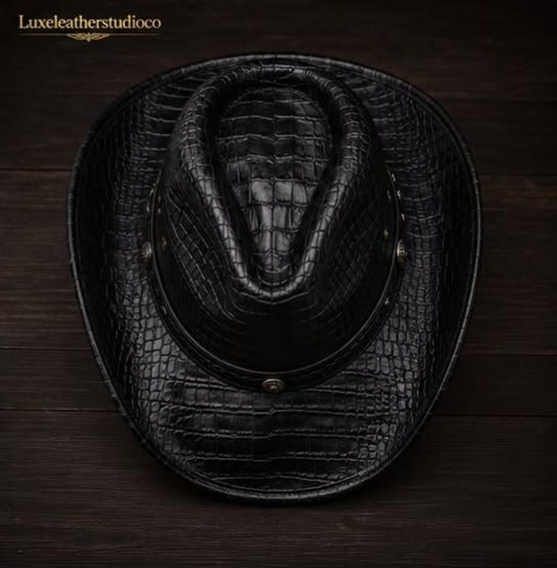 K&ouml;nnte beinhalten: Ein schwarzer Cowboyhut mit einer strukturierten Krokodilleder-Optik auf der gesamten Oberfl&auml;che. Der Hut hat ein dunkles Band um die Krone, das mit kleinen, runden, silberfarbenen Metallnieten verziert ist. Der Text Luxeleatherstudioco ist sichtbar.