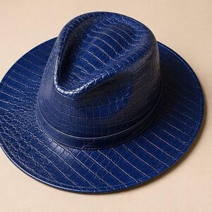 May include: A deep blue fedora hat featuring a glossy, embossed crocodile skin texture across the entire surface. The hat has a structured crown with a central crease and a wide, flat brim, finished with a matching blue band around the base.