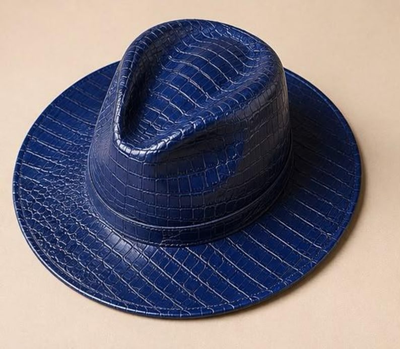 May include: A deep blue fedora hat featuring a glossy, embossed crocodile skin texture across the entire surface. The hat has a structured crown with a central crease and a wide, flat brim, finished with a matching blue band around the base.