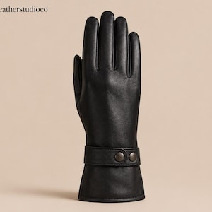May include: A black leather glove featuring a smooth finish and a wrist strap secured with two metallic snap buttons. The glove displays a classic five-finger design with visible stitching along the fingers and a clean, straight-cut cuff edge.