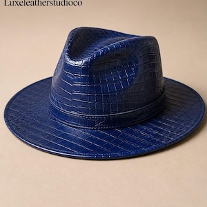May include: A deep blue fedora hat featuring a textured crocodile skin pattern across the crown and wide brim. The hat includes a matching blue band around the base of the crown. The text Luxeleatherstudioco is visible in the upper left corner.