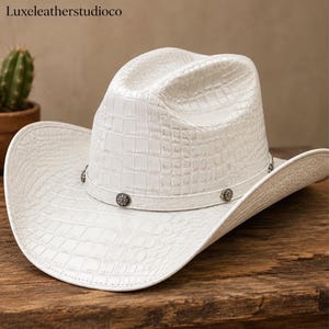 Op de afbeelding: Een witte cowboyhoed met een textuur van krokodillenleer over de kroon en de rand. De hoed is voorzien van een bijpassende witte band versierd met kleine, ronde, zilverkleurige metalen studs met een bloemmotief rond de basis.