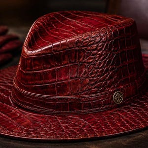 Può includere: Un cappello fedora rosso scuro con un motivo testurizzato in pelle di coccodrillo e un paio di guanti in pelle abbinati. Il cappello presenta una corona strutturata, una tesa larga e un emblema metallico circolare sulla fascia. Il testo luxeleatherstudioco è visibile.