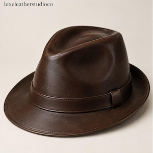 May include: A dark brown leather fedora hat with a creased crown and a matching leather band around the base. The text 'luxeleatherstudioco' is visible in the upper left corner. The hat features a smooth texture and a wide, curved brim.