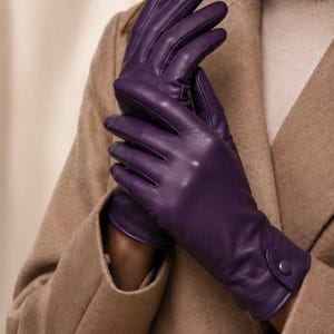 May include: A pair of deep purple leather gloves featuring a smooth finish, stitched detailing along the fingers, and a small button closure at the wrist. The text Luxeleatherstudioco is visible in the upper left corner of the image.