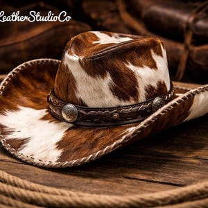 May include: A cowboy hat featuring a brown and white cowhide pattern with a dark brown leather hat band adorned with silver-toned metal conchos. The brim edge is finished with a braided leather trim. The text LuxeLeatherStudioCo is visible.