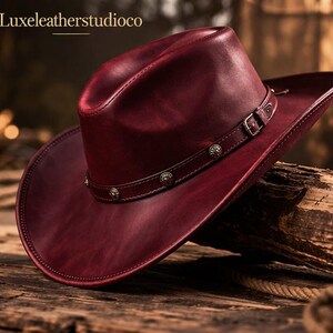 May include: A burgundy leather cowboy hat with a wide brim and a decorative band. The hat features a buckle and metal studs. The text "Luxeleatherstudioco" is at the top.