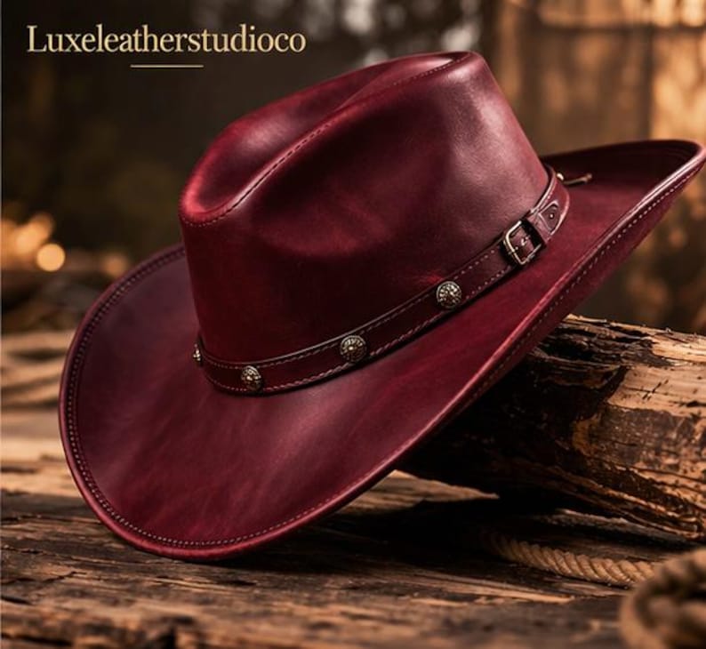 May include: A burgundy leather cowboy hat with a wide brim and a decorative band. The hat features a buckle and metal studs. The text "Luxeleatherstudioco" is at the top.