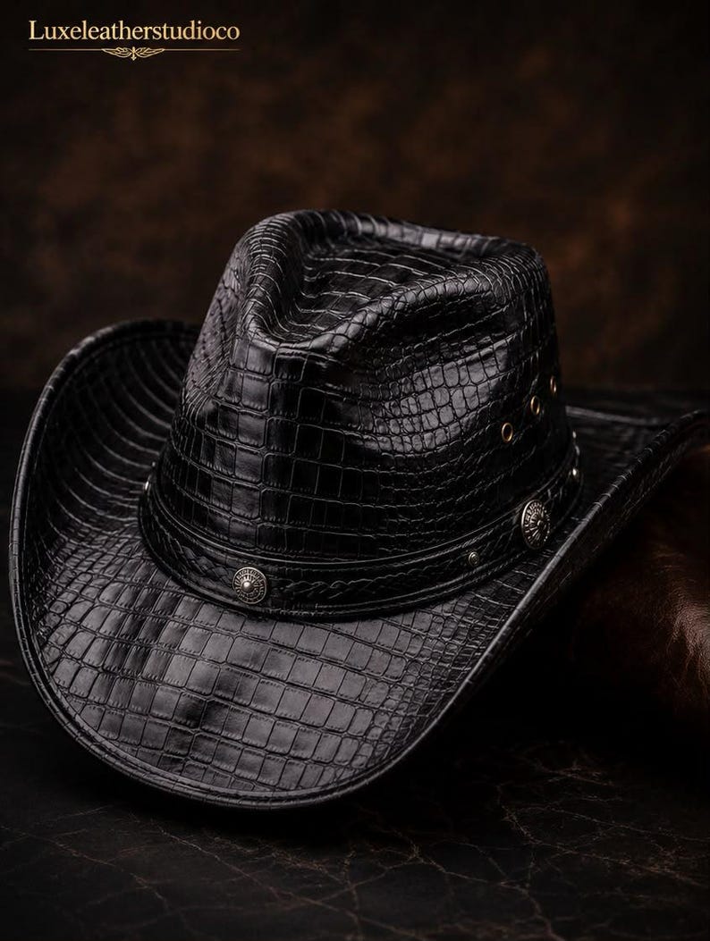 K&ouml;nnte beinhalten: Ein schwarzer Cowboyhut mit einer strukturierten Krokodilleder-Optik. Der Hut verf&uuml;gt &uuml;ber ein dekoratives Band, das mit runden, silberfarbenen Metallnieten und &Ouml;sen an der Seite verziert ist. Der Text Luxeleatherstudioco ist oben links zu sehen.