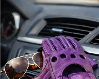 May include: A pair of purple leather driving gloves with cut-out details and a pair of gold-rimmed sunglasses rest on a car dashboard. The gloves are a vibrant shade of purple, and the sunglasses have brown lenses.
