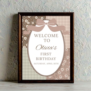Könnte beinhalten: Ein gerahmtes Schild mit einem dunkelbraunen Rahmen. Der Text lautet "WELCOME TO Olivia's FIRST BIRTHDAY" in einer dekorativen Schriftart. Der Hintergrund ist ein Patchwork aus Blumen- und Vichymustern in Braun und Creme. Oben befindet sich eine dekorative Schleife.