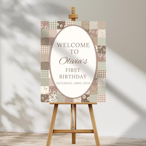 Könnte beinhalten: Eine Holzstaffelei zeigt ein Willkommensschild für einen ersten Geburtstag. Das Schild hat einen ovalen Rahmen mit dem Text "Welcome to Olivia's First Birthday" und einen Patchwork-Hintergrund in Braun-, Rosa- und Grüntönen. Das Datum ist Samstag, 18. April.