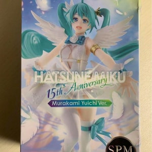 Puede incluir: Caja SEGA con una figura de Hatsune Miku que celebra el 15 aniversario. La figura tiene cabello turquesa y viste un atuendo blanco y azul claro con alas de ángel. La caja tiene el texto "Hatsune Miku" y "SPM Super Premium".