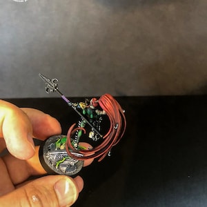 以下が含まれることがあります： 長い赤い髪をなびかせた戦士のミニチュアフィギュア。紫色の柄の槍を持っている。フィギュアは緑と茶色のディテールが施された丸い台座の上に立っている。台座には「KING」の文字が書かれている。