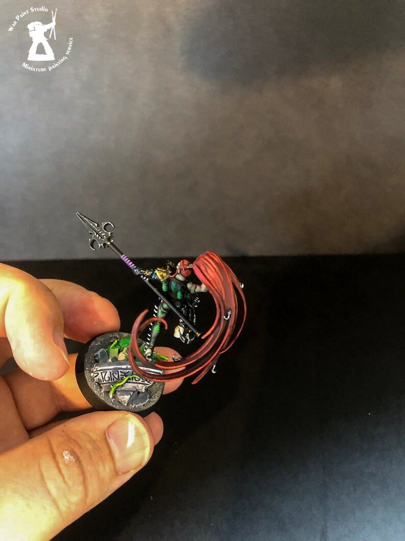 以下が含まれることがあります： 長い赤い髪をなびかせた戦士のミニチュアフィギュア。紫色の柄の槍を持っている。フィギュアは緑と茶色のディテールが施された丸い台座の上に立っている。台座には「KING」の文字が書かれている。