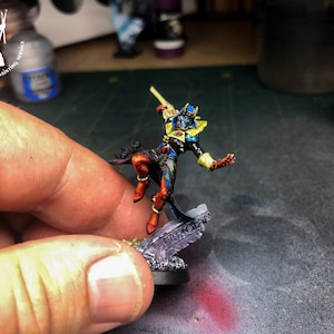 May include: A blue and red painted miniature figure of a warrior holding a staff, standing on a gray stone base. The figure is wearing a blue and gold helmet and a red and gold robe.