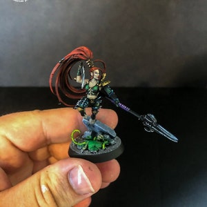 以下が含まれることがあります： 赤い髪の女性戦士の緑と黒のミニチュアフィギュア。彼女は片手に剣、もう片手に槍を持っています。フィギュアは、頭蓋骨と緑色の生き物が置かれた台座の上に立っています。