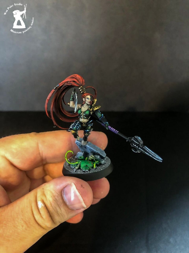 以下が含まれることがあります： 赤い髪の女性戦士の緑と黒のミニチュアフィギュア。彼女は片手に剣、もう片手に槍を持っています。フィギュアは、頭蓋骨と緑色の生き物が置かれた台座の上に立っています。