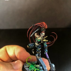 以下が含まれることがあります： 赤い長い髪をした緑と黒の女性戦士のミニチュアフィギュア。彼女は槍と短剣を持っています。フィギュアは緑と灰色の岩の形成をした黒いベースの上に立っています。