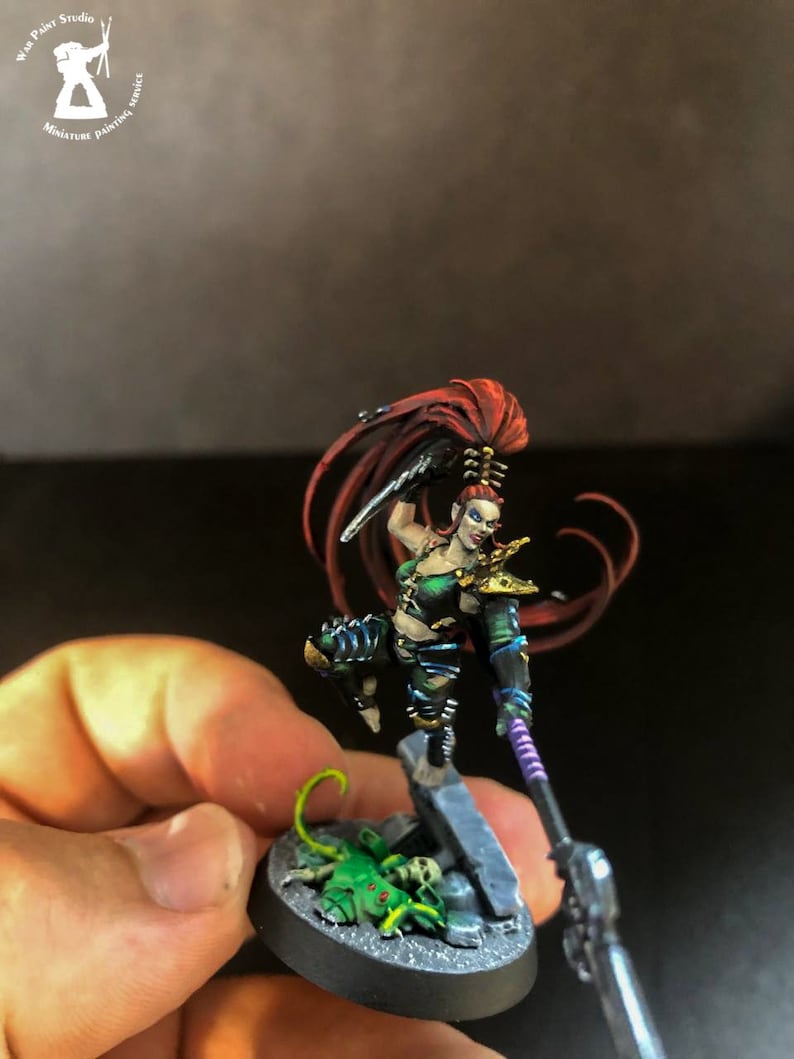 以下が含まれることがあります： 赤い長い髪をした緑と黒の女性戦士のミニチュアフィギュア。彼女は槍と短剣を持っています。フィギュアは緑と灰色の岩の形成をした黒いベースの上に立っています。