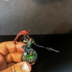 以下が含まれることがあります： 長い赤い髪の女性戦士のミニチュアフィギュア。緑と黒の鎧を着て、槍を持っています。フィギュアは緑と黒のデザインの丸い台座の上に立っています。