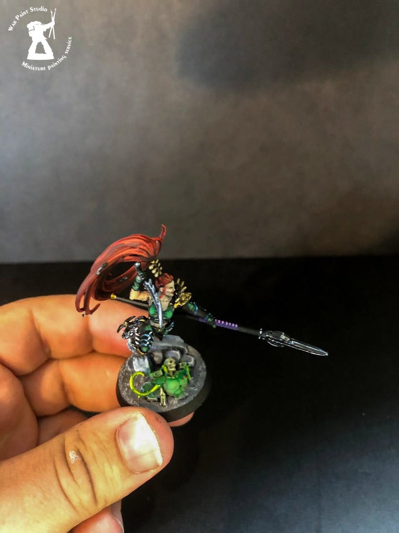 以下が含まれることがあります： 長い赤い髪の女性戦士のミニチュアフィギュア。緑と黒の鎧を着て、槍を持っています。フィギュアは緑と黒のデザインの丸い台座の上に立っています。