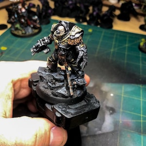 May include: A black and gold painted miniature figure of a space marine with a large gun and a sword. The figure is standing on a rocky base.