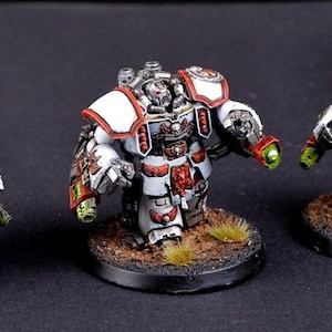 May include: Three miniature figurines of armored figures, painted in white, red, and silver. Each figure stands on a circular base with a textured, brown surface. The figures have detailed armor and weaponry, with green accents.