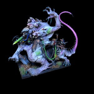 May include: A multi-headed, pale purple creature figurine with a long, curved, bright pink tail. The creature features exposed muscle textures, green glowing accents, and a small rider figure mounted on its back, all set on a rectangular base.