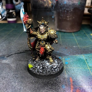 Puede incluir: Una figura en miniatura pintada de oro de un guerrero que lleva una corona y sostiene un hacha grande. La figura está de pie sobre una base negra con un pequeño parche de hierba verde.