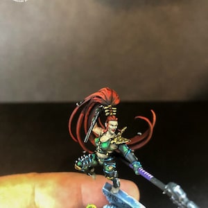 以下が含まれることがあります： 赤い長い髪をした女性戦士のミニチュアフィギュア。緑と黒の鎧を着て、槍と短剣を持っている。頭蓋骨と緑色の生き物が置かれた台座の上に立っている。