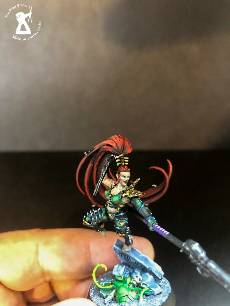 以下が含まれることがあります： 赤い長い髪をした女性戦士のミニチュアフィギュア。緑と黒の鎧を着て、槍と短剣を持っている。頭蓋骨と緑色の生き物が置かれた台座の上に立っている。