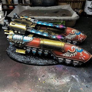 Könnte beinhalten: Zwei gold- und rot lackierte Miniaturpanzer mit blau-weißen Adler-Insignien. Die Panzer haben ein futuristisches Design mit mehreren Kanonen und Rohren.