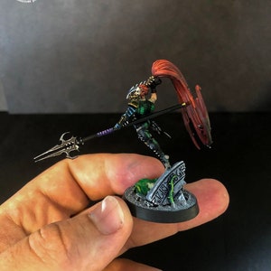 以下が含まれることがあります： 槍と赤い布を持った戦士のミニチュアフィギュア。フィギュアは緑、黒、銀で塗装されています。フィギュアは緑と銀の墓石のある黒い台座の上に立っています。