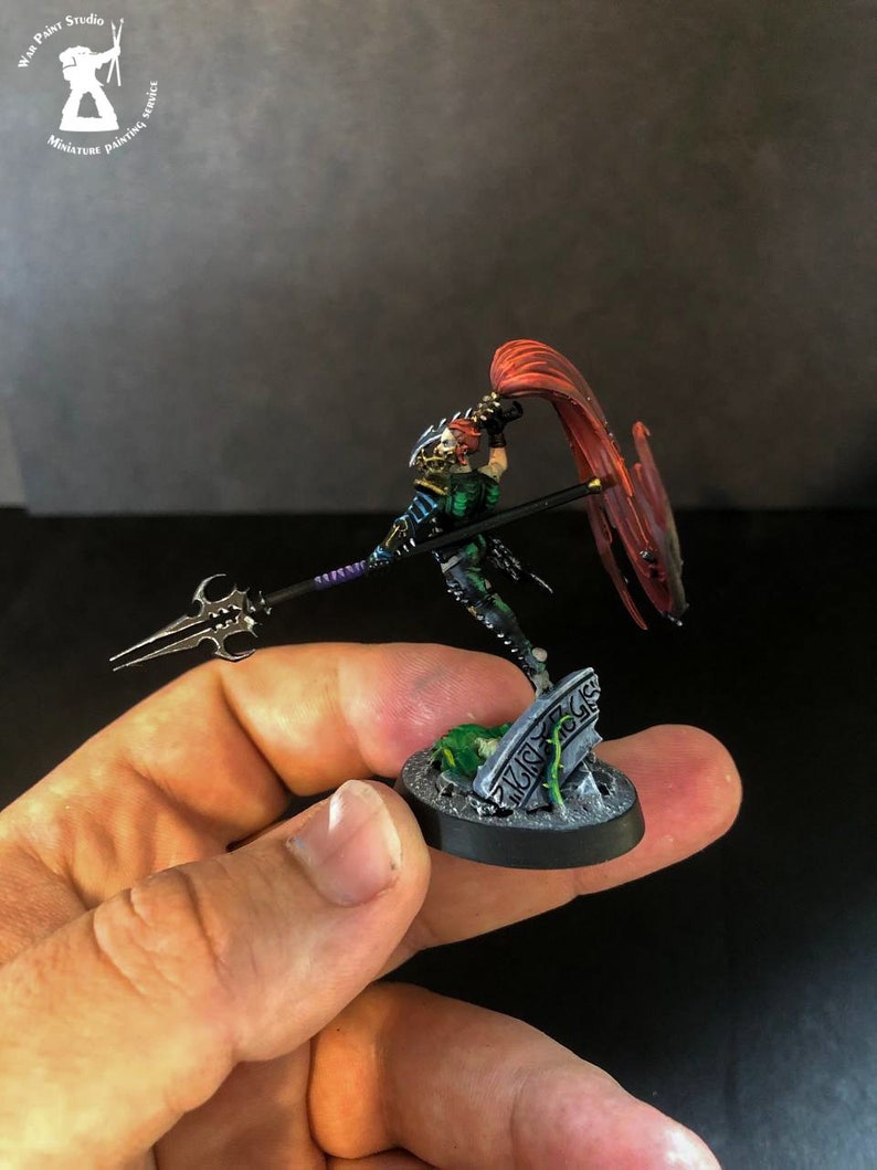 以下が含まれることがあります： 槍と赤い布を持った戦士のミニチュアフィギュア。フィギュアは緑、黒、銀で塗装されています。フィギュアは緑と銀の墓石のある黒い台座の上に立っています。