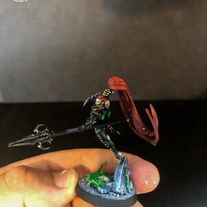 以下が含まれることがあります： 赤いマントをつけた黒と緑のミニチュアフィギュアで、紫色の柄の槍を持っています。フィギュアは、緑と灰色の墓石のある円形の台座の上に立っています。