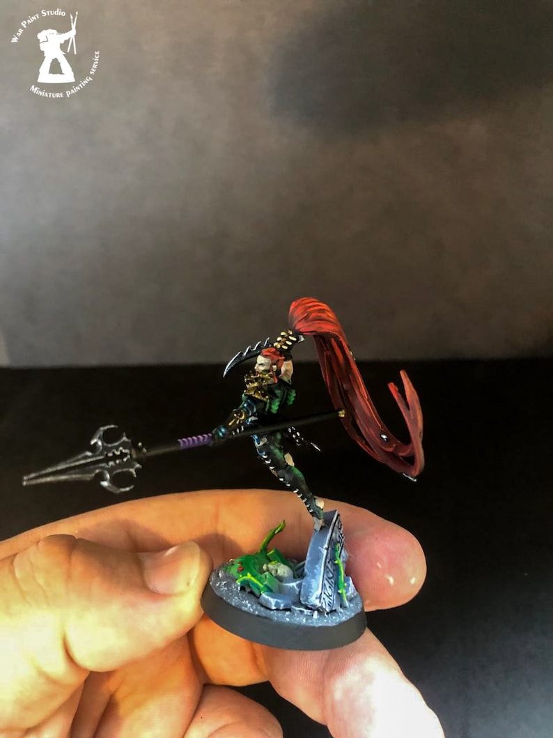 以下が含まれることがあります： 赤いマントをつけた黒と緑のミニチュアフィギュアで、紫色の柄の槍を持っています。フィギュアは、緑と灰色の墓石のある円形の台座の上に立っています。