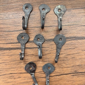 Puede incluir: Siete ganchos de metal para marcos de fotos de estilo antiguo. Cada gancho es de color gris oscuro con una parte superior en forma de ojo de cerradura y un gancho curvo. Los ganchos están dispuestos sobre una superficie de madera.