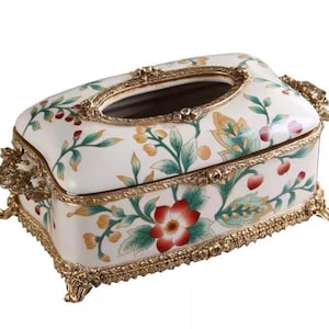 May include: An ornate, rectangular tissue box with a white background and floral design. The box features a gold-colored trim and feet, with a decorative opening on top. The floral pattern includes red, green, and yellow accents.