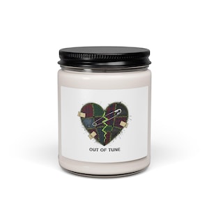 May include: A white candle jar with a black lid. The label features a heart design made of patchwork fabric, a safety pin, and the text "OUT OF TUNE". The heart is split with a jagged green line and has small beige patches.