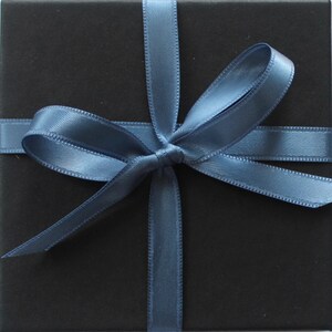 Pu&ograve; includere: Una scatola regalo quadrata nera avvolta da un nastro di raso blu ardesia liscio legato in un fiocco centrale. Il nastro si incrocia orizzontalmente e verticalmente sulla superficie nera opaca della scatola, creando una presentazione ordinata.