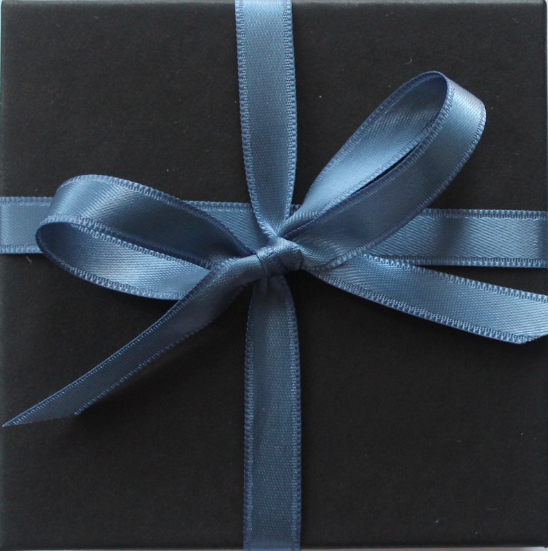 Pu&ograve; includere: Una scatola regalo quadrata nera avvolta da un nastro di raso blu ardesia liscio legato in un fiocco centrale. Il nastro si incrocia orizzontalmente e verticalmente sulla superficie nera opaca della scatola, creando una presentazione ordinata.