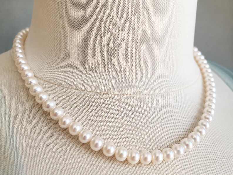 Pu&ograve; includere: Una collana a filo singolo composta da perle rotonde e uniformi di colore bianco sporco. Le perle presentano una superficie liscia e lucente, infilate in una linea continua che poggia su un busto espositore in tessuto di colore neutro.