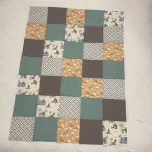 May include: A patchwork quilt with a grid design, featuring squares in different colours and patterns. The squares include solid grey, teal, and patterned fabrics with cartoon illustrations and plaid designs. The quilt is likely handmade.
