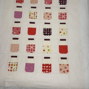 May include: A patchwork quilt with a repeating design of jar-shaped blocks. Each jar has a white top and a body filled with patterns such as hearts, strawberries, and polka dots. The background is white with small, evenly spaced dots.