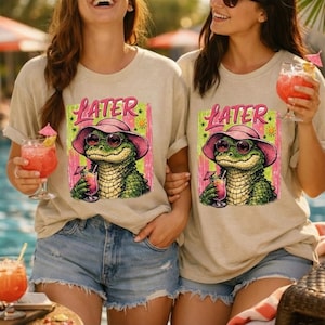 Könnte beinhalten: Zwei beige T-Shirts mit einer Grafik eines Alligators mit Sonnenbrille und rosa Hut, der einen Cocktail hält. Das Wort "LATER" steht über dem Alligator. Die Shirts werden mit Jeansshorts kombiniert.